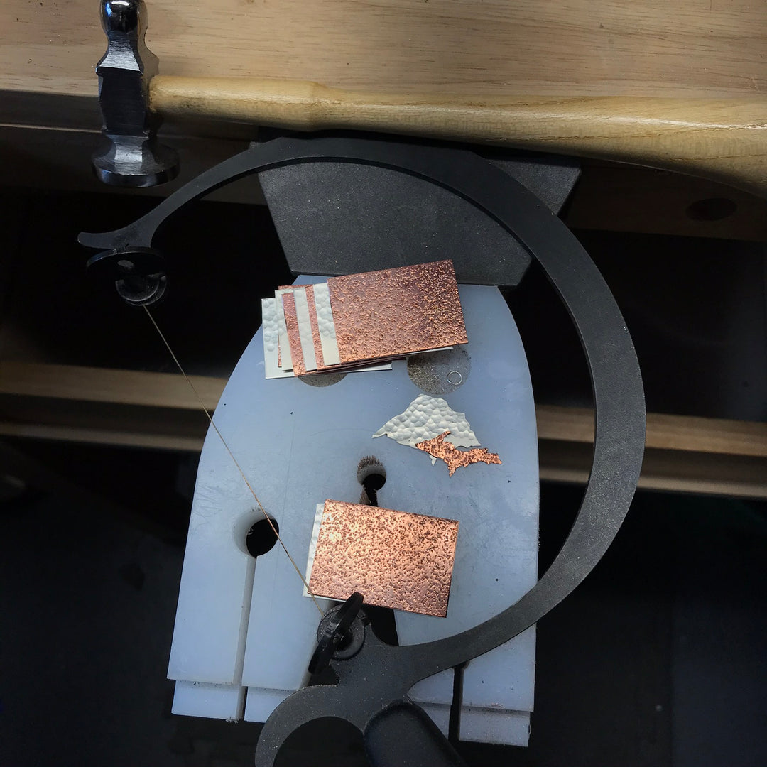 Metalsmith's bench pin with a jeweler's saw and pieces of textured silver and copper with a silver Lake Superior and a copper Upper Peninsula sawn out.