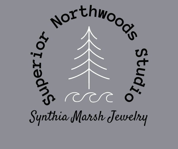 A simple pine tree shape with three waves underneath in white with a circular Superior Northwoods Studio placed around the tree and the words Synthia Marsh Jewelry underneath.