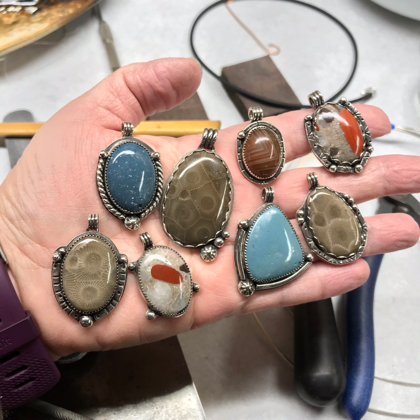 Hand holding various Lake Superior area and Michigan (Upper Peninsula) stone pendants including Petoskey Stones, Leland Blue, Lake Superior Agates and Pudding Stones in Sterling Silver settings made by Synthia Marsh Jewelry