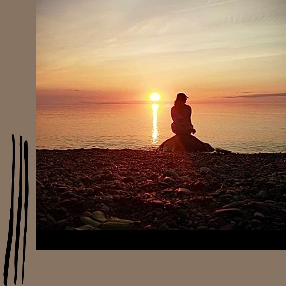 Silhouette of Synthia Marsh sitting on a large rock on the calm edge of Lake Superior with the setting sun.