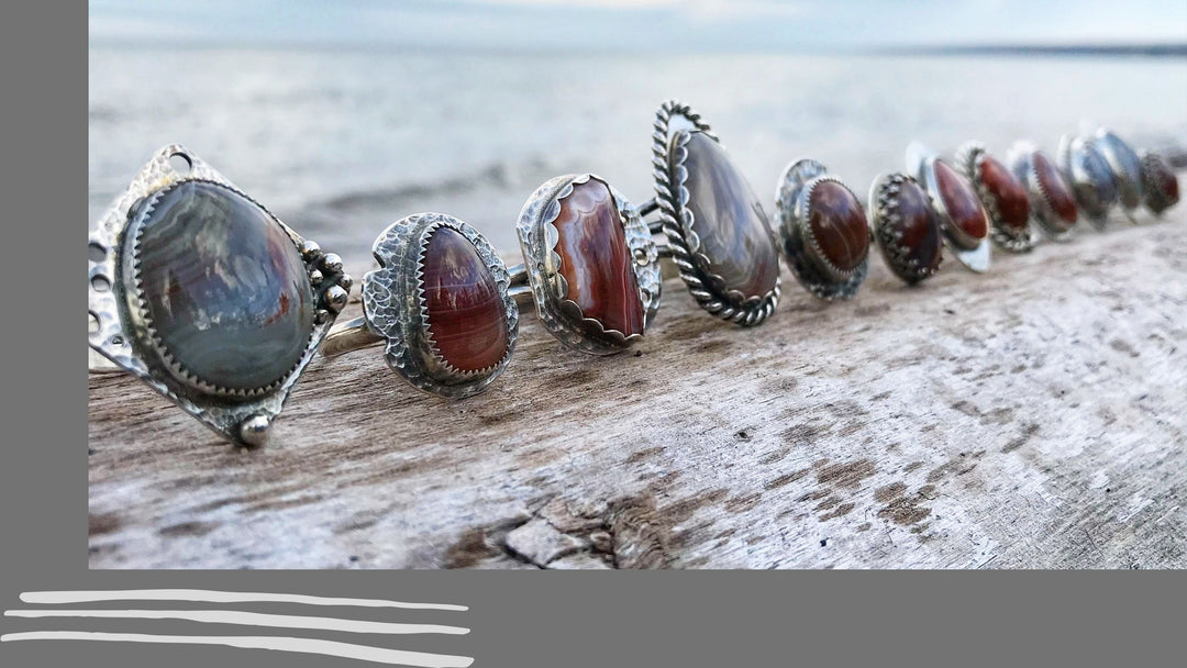 Twelve sterling silver and Lake Superior Agate rings lined up on a piece of driftwood at the shore of Lake Superior.