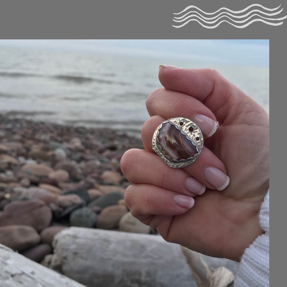A beautiful Lake Superior Agate ring modeled on a hand with the rocky shoreline of Lake Superior in the background.