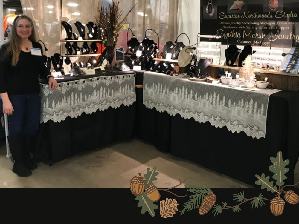 Synthia Marsh standing in her jewelry booth set-up with two tables of various types of jewelry displayed on a black and ivory lace display.