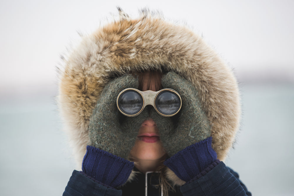 Woman with winter fake-fur lined hood, woolen mittens and looking straight at camera through a pair of binoculars.